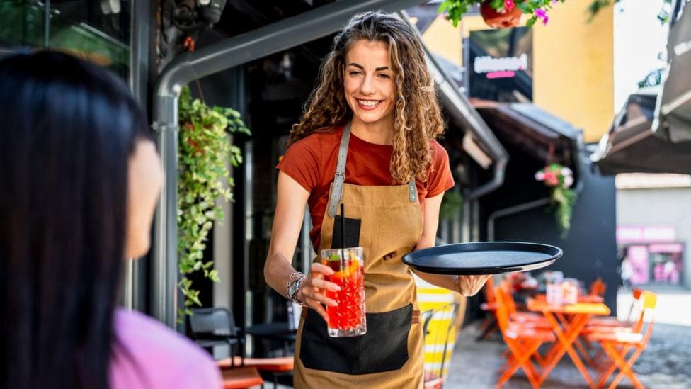 A woman working in a bar serves a cocktail drink to another woman at a table.