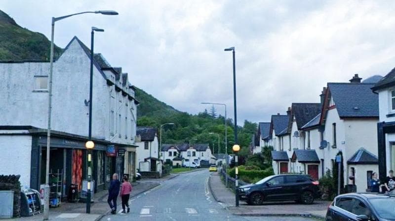 Street view of Leven Road in Kinlochleven. Houses sit along one side of the street and a small supermarket is on the other. There is a zebra crossing and two people cross the street.