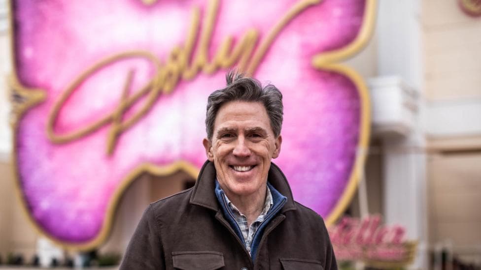 Rob Brydon smiling at the camera. He had brown hair and wearing a brown jacket. There's a big pink butterfly sign behind him with Dolly written in yellow.