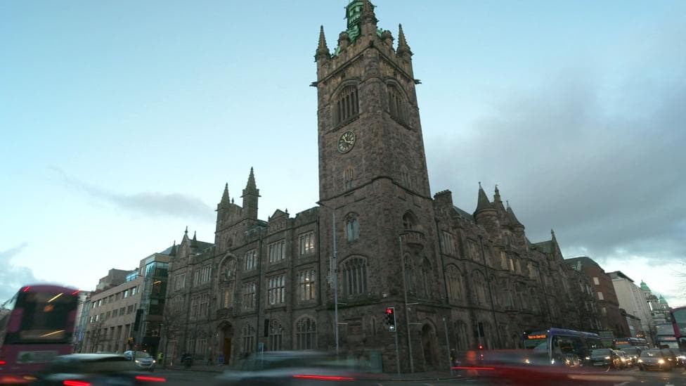 A large church building in Belfast city centre shows, stained glass windows and a bell tower, bearing a gold and black clock. Behind the building is a gloomy, cloudy sky. Traffic is blurred as it passes the camera.