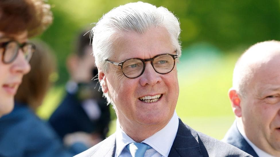 Malcolm Offord had short white hair and wears glasses and a dark suit. He smiles looking off to the side and appears to be outside with a man on either side of him