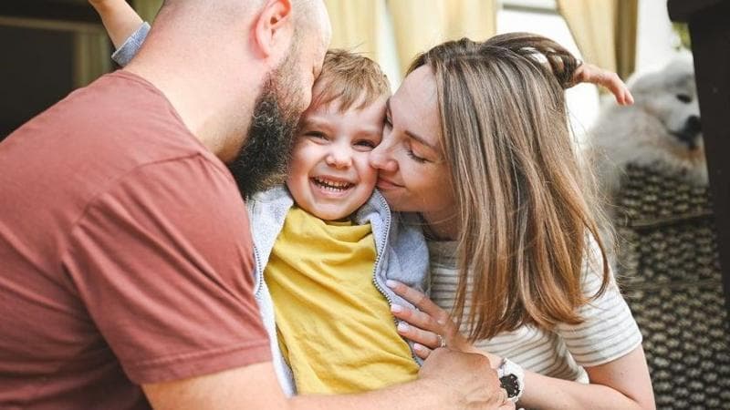 A young blonde child wearing a yellow t-shirt is hugged by a man wearing a red t-shirt and a woman wearing a beige striped t-shirt.