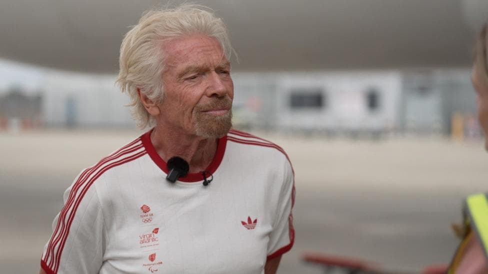 Richard Branson wearing a white t-shirt with the Team GB, Virgin Atlantic, and Paralympics GB logos in the right side of the chest with red lettering, an Adidas logo on the left side of the chest in red, and three red stripes on each arm. He is standing on the runway of Heathrow Airport. Behind him are a Virgin Atlantic plane and the terminal buildings in soft focus.