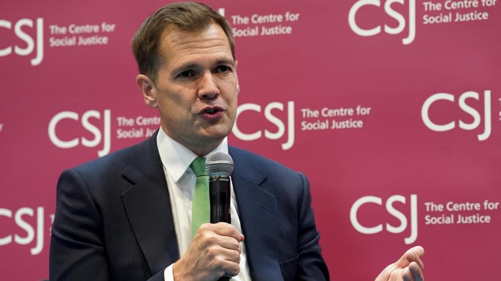 Robert Jenrick holding a microphone wearing a dark suit and a green tie. He is sitting in front of a branded sign for the Centre for Social Justice