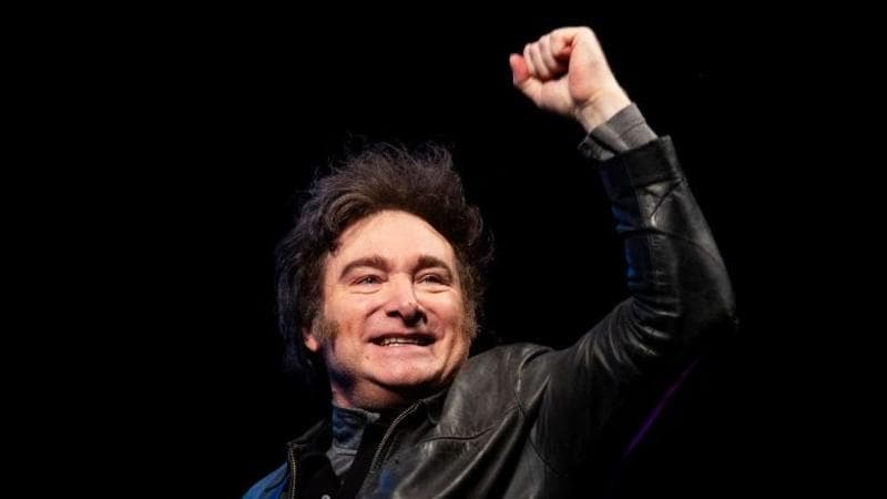 Argentine President Javier Milei, wearing a leather jacket, stands smiling with a closed fist raised against a black backdrop.