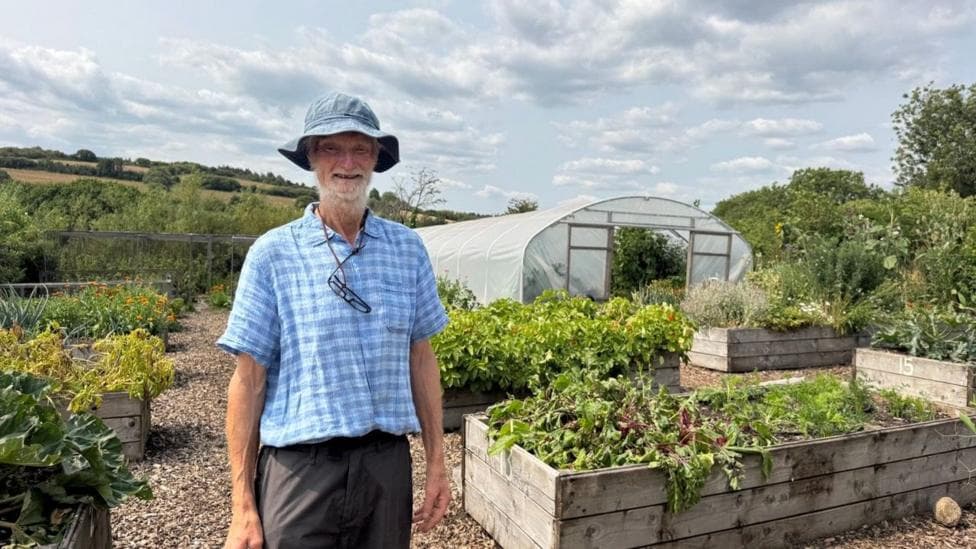 Man in a blue checkered shirt and a bucket hat, standing off the the left of a planter at a farm in Dartmoor. In the background there is a poly-tunnel greenhouse and rolling hills.