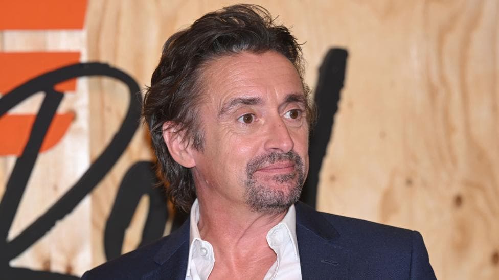 Richard Hammond is photographed from the shoulders up, standing in front of a promotional sign for the TV series 'The Grand Tour'. His face is facing slightly to the side and he is not looking at the camera. He is wearing a black suit jacket and a white shirt, with the top button undone.