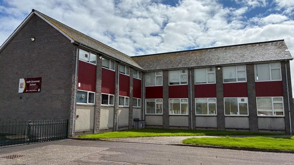 An L-shaped grey school building with a maroon sign on the side, reading 'Keith Grammar School'
