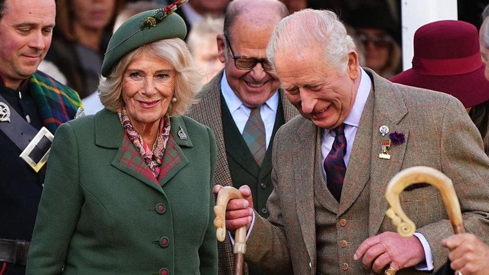 Queen Camilla, wearing a green blazer and a green hat, next to King Charles who is wearing a tweed three-piece suit. They are surrounded by officials and are in high spirits at the Braemar Gathering