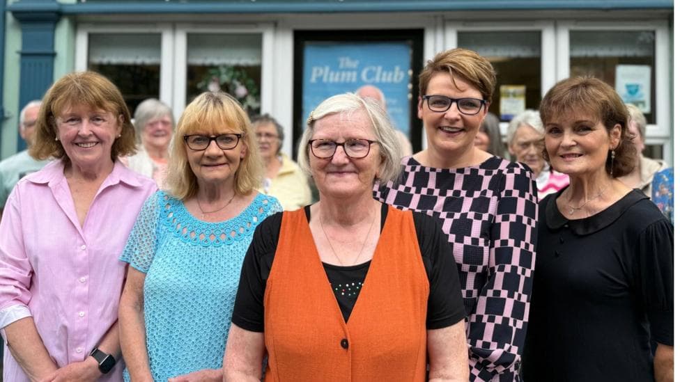 Five women standing smiling at camera.
Women one left is wearing a pink shirt, second left is blue top, Anne Bradley is in the middle wearing orange dress and black t-shirt, beside her on right is woman with chequered top and then last woman is wearing a black top.