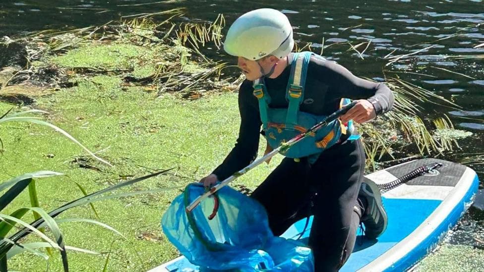 One of the litter pickers is kneeling on a surfboard while picking rubbish out of water with a grabber and putting it in a blue bag. He is in a wetsuit and has on a life-vest and helmet.