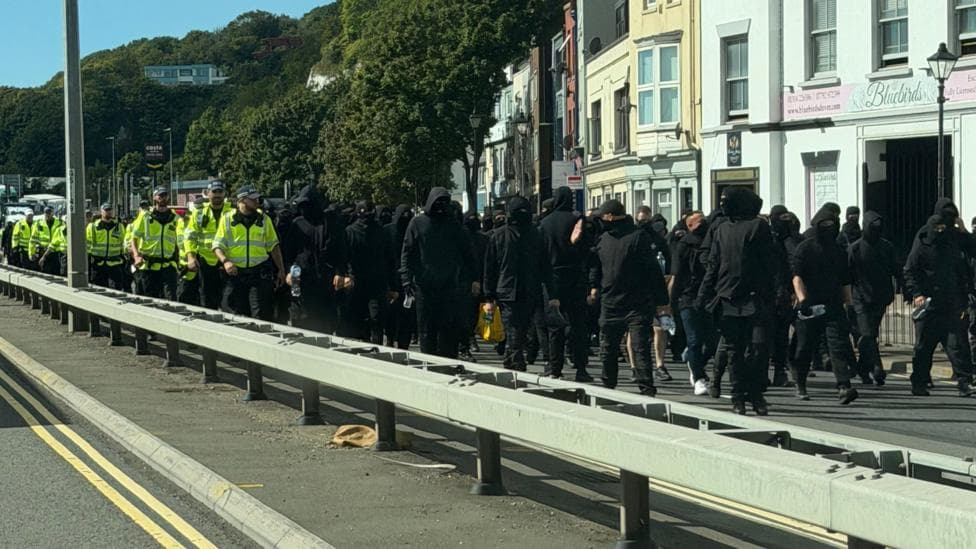 Dozens of men dressed in black and wearing face coverings, being escorted down a dual carriageway in Dover by uniformed police officers.