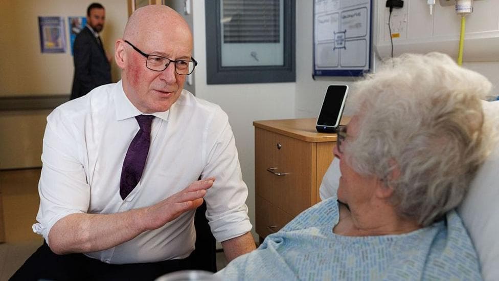 John Swinney talking to a female hospital patient who is in bed.