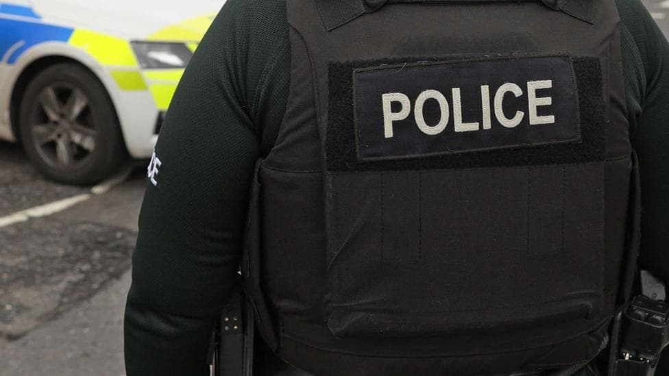 A close-up image of a the back of a police officer's black protection vest as they stand near a police car