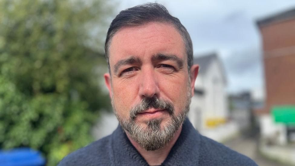 Paul Doherty has short dark hair and a black and grey beard. He is wearing a navy jumper cardigan.