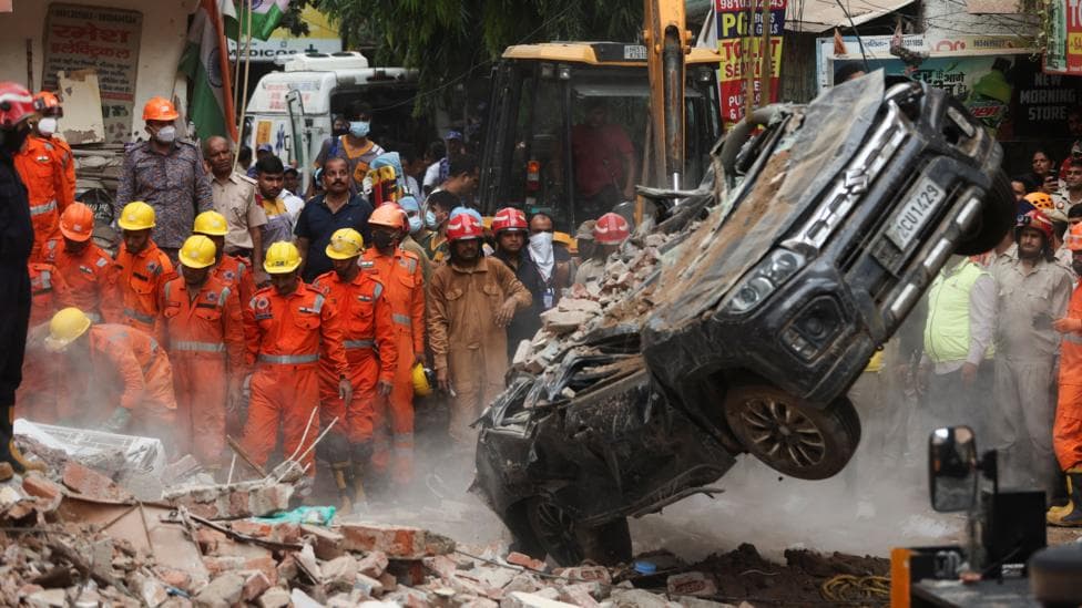 National Disaster Response Force (NDRF) personnel look on at a building collapse in Delhi, while a crushed car is pulled by a digger from the rubble.