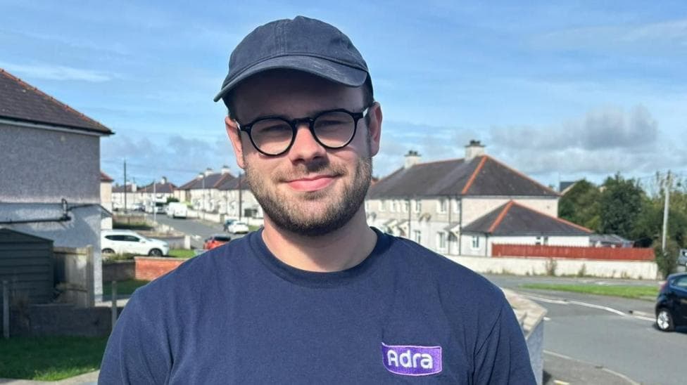 A picture of a man wearing a blue cap and a blue tshirt with glasses. He's smiling at the camera and standing in a housing estate