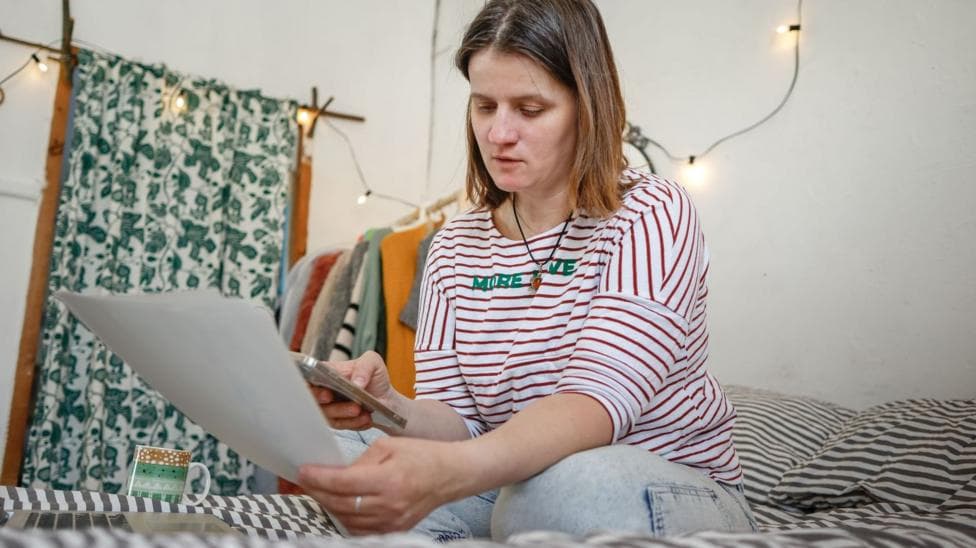 Woman in a red and white striped top sits on a bed looking at a letter and a phone with a rack of clothes and a curtain beside her.