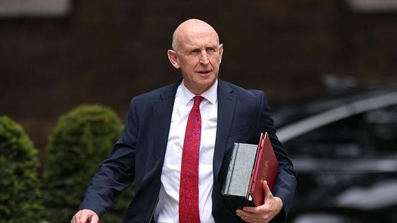 An image of John Healey from the waist up. He is walking and wearing a dark suit with a white shirt and red patterned tie. He is carrying a red folder. The background is blurred by you can see that he is outside from the outline of bushes.