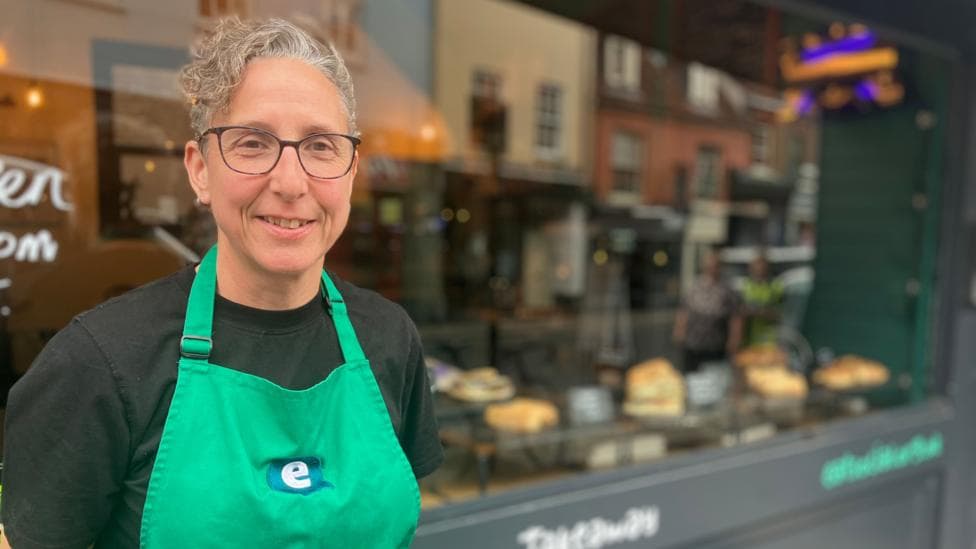 A woman wearing glasses ,a dark top and a green apron stands outside a café, which is out of focus behind her