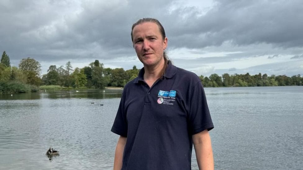 There is a man in a dark blue polo shirt which has two symbols on his chest pocket. The top symbol says "Shropshire Council" on a dark blue background. Below it, it says "Shropshire's Great Outdoors." He is stood in a lake and has a duck over to his right.