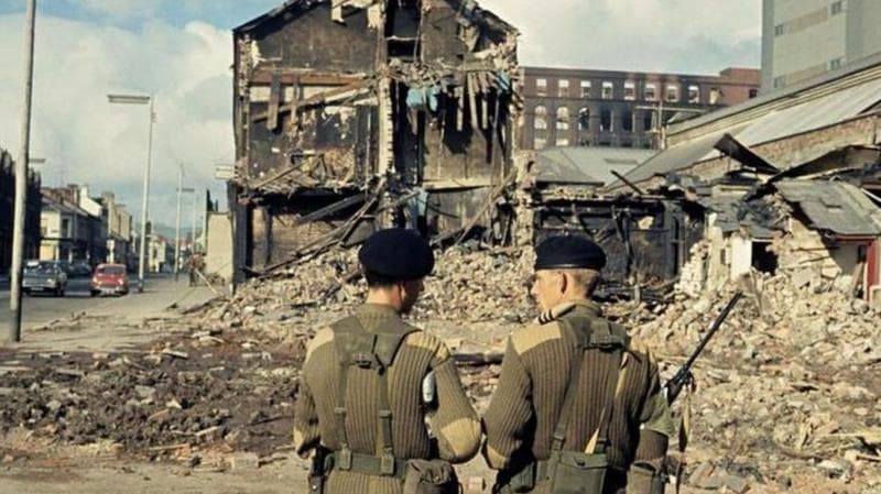 A historic, colourised image of two soldiers standing in front of a pile of rubble and a destroyed building.