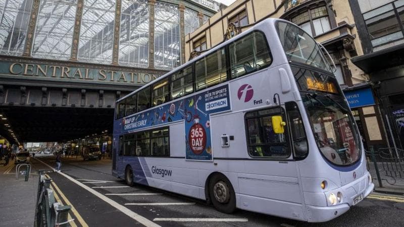A white First Bus double decker bus emerges from under the "Heilanman's umbrella", the bridge under Glasgow's main railway station. Behind it a huge green sign with gold lettering says "Central Station".There is ornate iron fencing on wither side of the road, which is marked up as a one way bus lane.