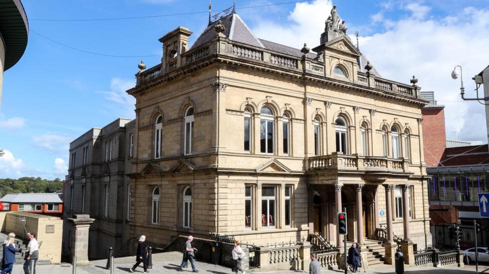 A grand old hall stands on a busy street in Londonderry, it has five storeys and a balcony can be seen to the front of the building, supported by columns. A number of pedestrians pass by.