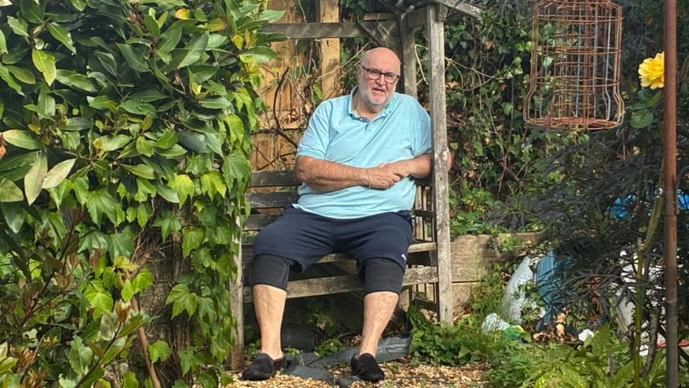 Phil Bond, 68 sat on a bench in his garden. He's wearing a light blue polo top and navy shorts, and slippers. He's bald with glasses. He's sat surrounded by greenery.