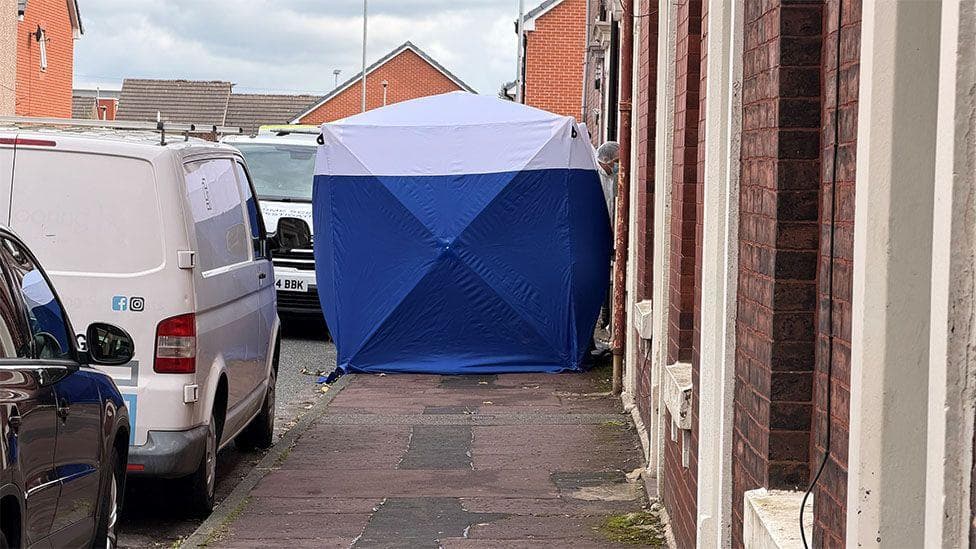Blue and white forensic tent on a residential pavement between parked vans and a row of terraced houses.