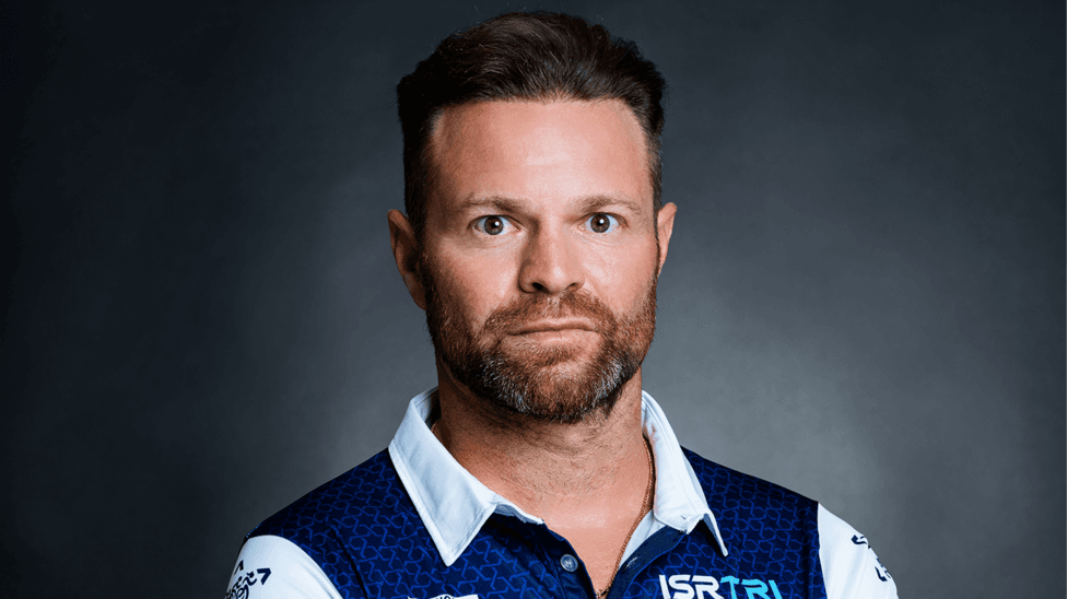 Lior Cohen stands against a dark background. He has short brown hair and beard and is wearing a blue and white Israeli Triathlon Association shirt
