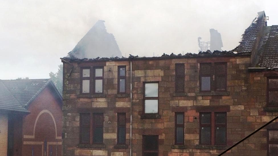 Burned-out tenement building which no longer has a roof. Three firefighters can be seen beside the building on the steps of a nearby flat