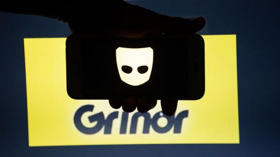 Grindr's app logo, displayed on a smartphone screen, is held in front of a yellow and black backdrop bearing the company's logotype.