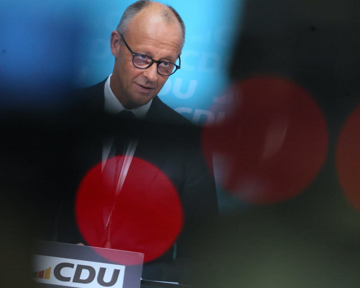 Friedrich Merz addresses a news conference in Berlin: he speaks from a lectern with the initials CDU on the front. Reflections and lighting project large red dots around him for a semi-abstract image.