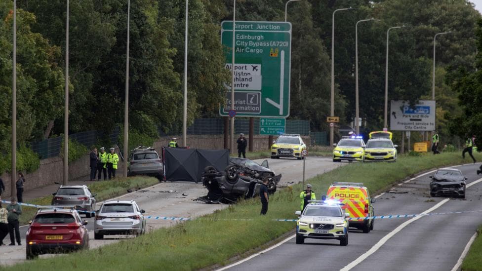 Aftermath of a road traffic collision, near Edinburgh Airport, featuring two badly damaged vehicles. One is on its roof and the other is on the opposite carriageway. The scene is cordoned off with police tape and officers in hi-viz jackets are on on the scene.