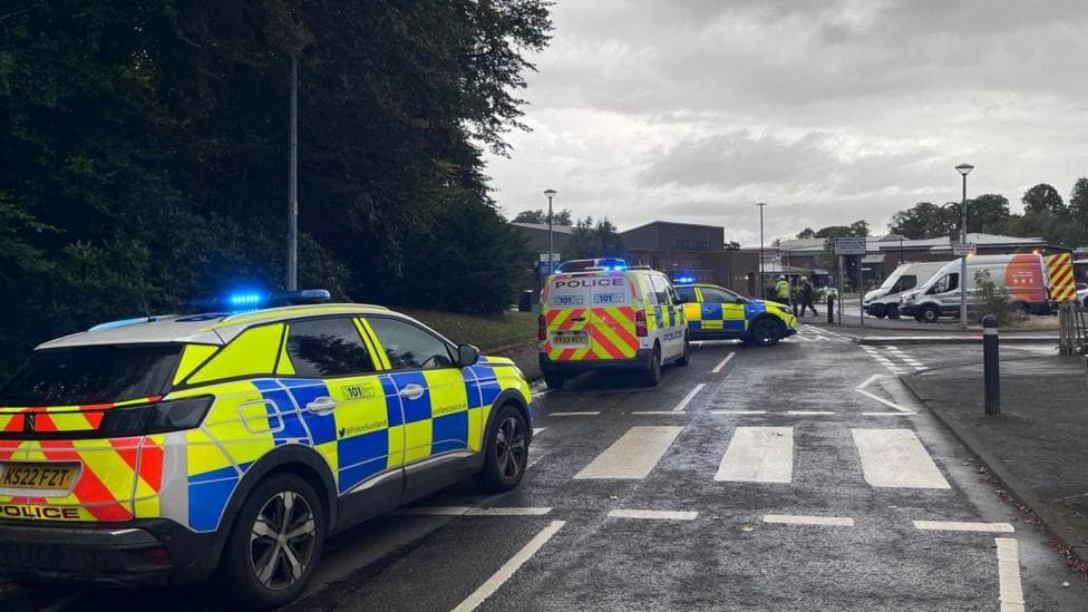 Three police vehicles are parked along a road with their flashing blue lights on. People in high vis gear can be seen in the distance