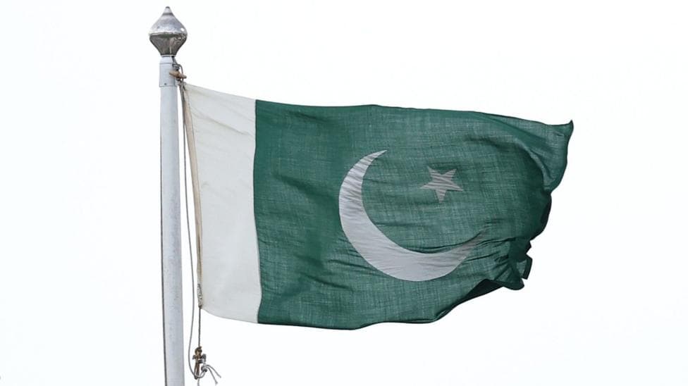 A Pakistan flag flying on a flag-pole. It is green with a white border down the left hand side, with a crescent moon and a five-pointed star in its centre.