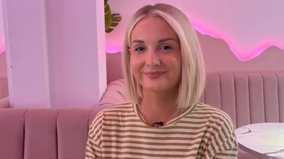 A picture of a woman with short blonde hair in a stripy top sitting in front of a pink background