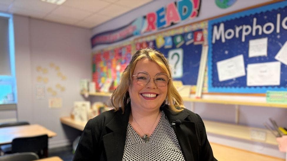 A smiling woman wearing glasses wearing a black jacket is sat in a school classroom