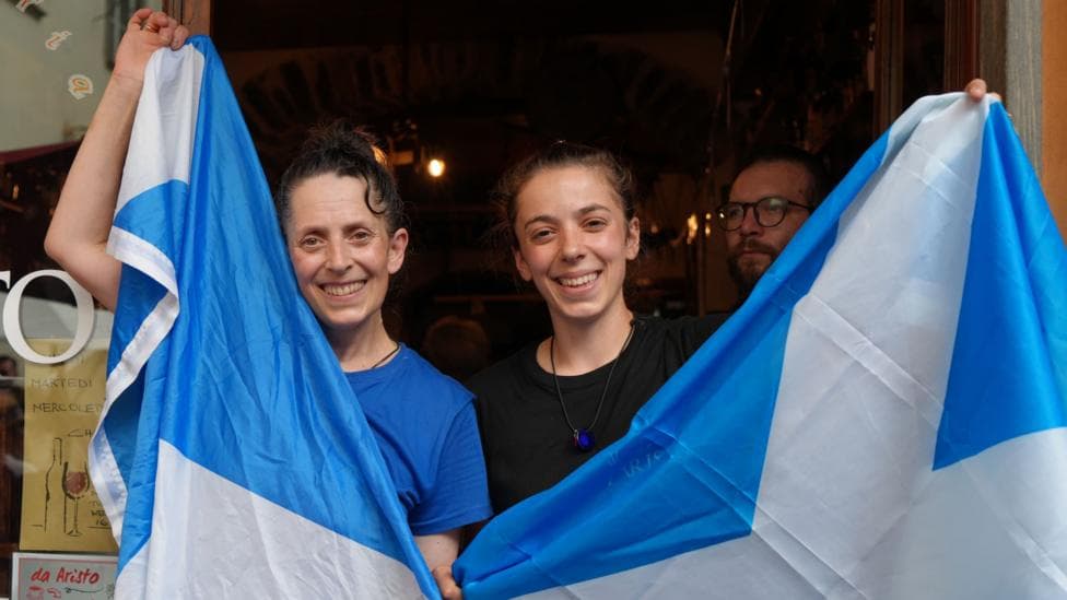 Two woman stand smiling behind Scotland flags