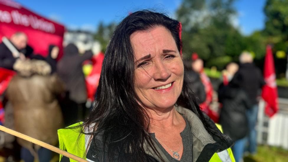 A woman stands by a road with a hi vis vest on. She is looking at the camera and her long dark hair is windswept. Behind her is a group of protesters holding large red flags with the Unite the union branding on them. Everyone is wearing rain coats but the sun is shining.