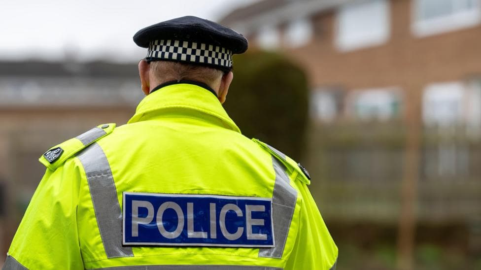 A policeman in a high-vis uniform jacket with 'POLICE' written on the back is facing away from the camera. He is wearing a police hat. There are buildings blurred in the background.
