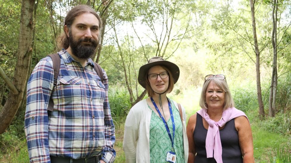 Alexander Jones is on the left of the picture. He is wearing a blue,red and white check shirt and has a long brown hair tied back. He also has a full beard. Georgina is in the middle, She is wearing a brown outdoor hat, a white cardigan over a green patterned dress. Councillor Patricia Hetherton is on the right of the image. She has a pink jumper tied by the sleeves around her neck over a black top. She has her glasses on the top of her head.