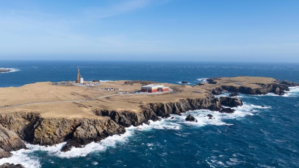 An aerial view of a long flat peninsula surrounded by dark blue sea. The coastline is rocky and the sky is blue. The island is brown and sparsely populated by several industrial buildings and a tall launch pad.
