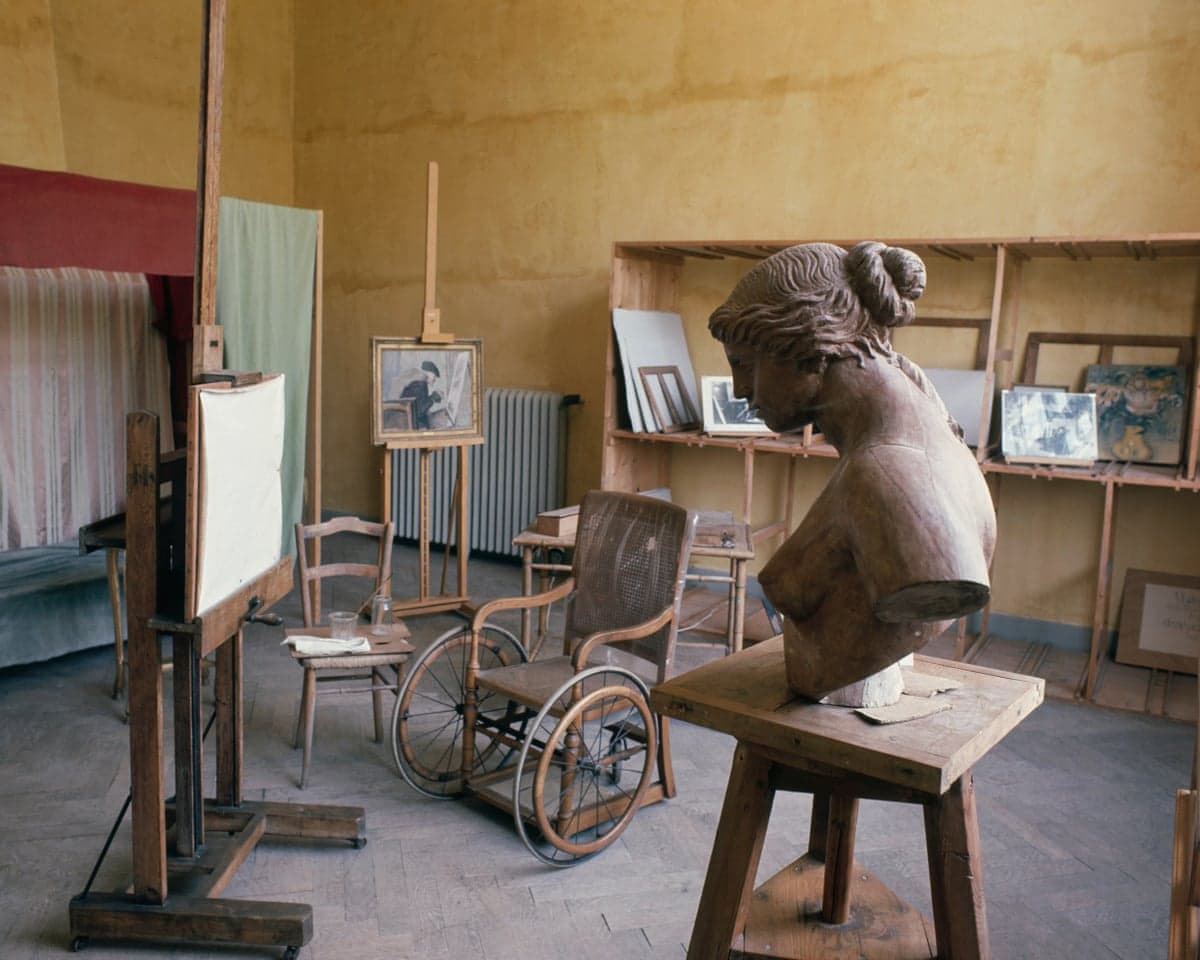 Renoir's studio with a sculpted bust, wheelchair, easels, and paintings on display