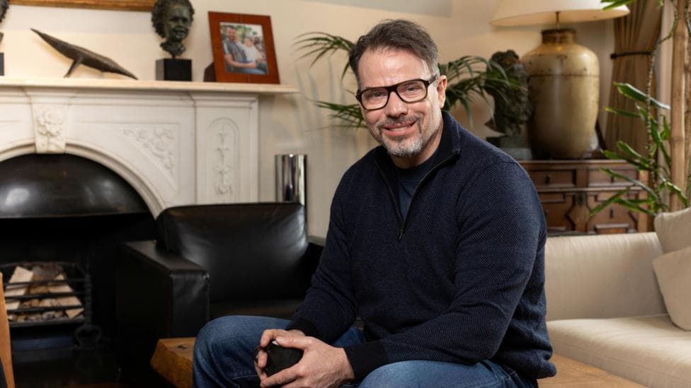 Chris Rokos sitting in a room wearing a dark blue jumper and blue jeans. In the background there is a fireplace, sofa and chest of drawers.