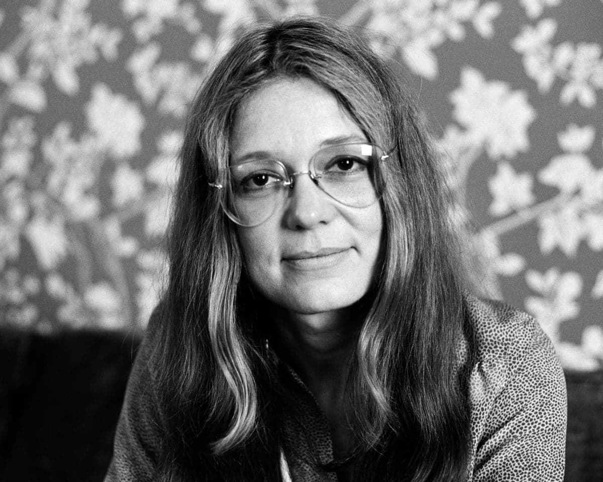 Black and white image of feminist writer and influencer Gloria Steinem, taken in 1977. She is looking into the camera, wearing a pair of wire-rimmed glasses, in front of a floral wall design