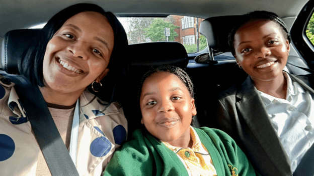 A smiling family sitting together in the back seat of a car. To the left is the mum, wearing a blue polka dot jacket. In the middle is a young girl in a green and yellow school uniform. On the right is an older girl in a white shirt and grey blazer.