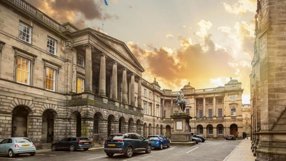 Historic stone government building with grand neoclassical columns surrounding a courtyard. Several parked cars and an equestrian statue stand in the foreground, while a dramatic golden sunset illuminates the clouds and architecture.