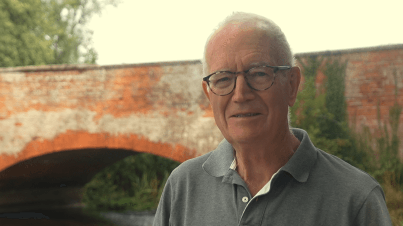 A man wearing a grey polo shirt and glasses. He is standing in front of a single-arched red brick bridge.